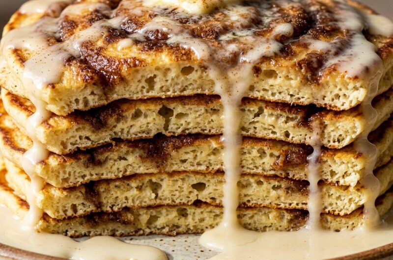 Ultra close-up macro of a thick fluffy stack of cinnamon roll pancakes with cascading cream cheese glaze and caramelized cinnamon swirls